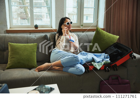 Happy, joyful young woman sitting at home, holding plane tickets, talking on phone and expressing excitement about upcoming trip Happy, joyful young woman sitting at home, holding plane tickets, talking on phone and expressing excitement about upcoming trip 111271761