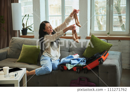 Young woman sitting on couch at home and packing her suitcase for travelling, picking outfits and putting shoes on luggage Young woman sitting on couch at home and packing her suitcase for travelling, picking outfits and putting shoes on luggage 111271762