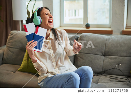 Happy and excited young woman listening to music in headphones, sitting on couch at home and showing passport with tickets. Happy and excited young woman listening to music in headphones, sitting on couch at home and showing passport with tickets. 111271766