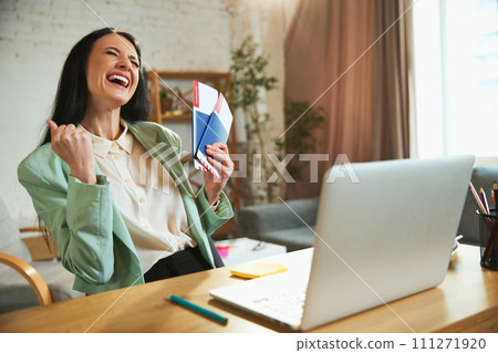 Happy, joyful young woman, employee sitting in office with laptop, holding plane tickets and showing excitement about upcoming trip 111271920