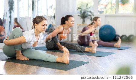 Concentrated young girl making stretching legs with group of active adult people sitting on yoga mat and doing exercise yoga 111272866