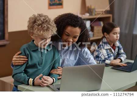 Portrait of smiling young Black teacher helping little boy using computer in school classroom copy space 111274794