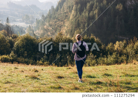 30s Girl with backpack on mountain peak with green grass looking at beautiful mountain valley in fog at sunset in summer. Landscape with sporty young woman, rear view. Full body 30s Girl with backpack on mountain peak with green grass looking at beautiful mountain valley in fog at sunset in summer. Landscape with sporty young woman, rear view. Full body 111275294