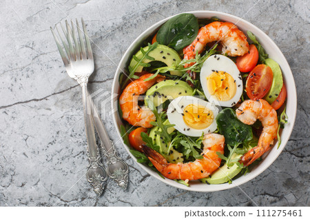 Delicious serving salad with shrimp, avocado, arugula, spinach, tomatoes, sesame seeds and boiled eggs close-up on a bowl. Horizontal top view 111275461