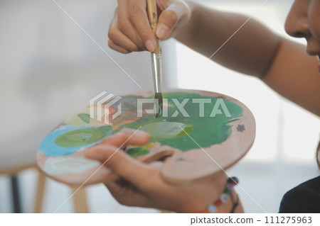 Cropped image of female artist standing in front of an easel and dipping brush into color palette 111275963