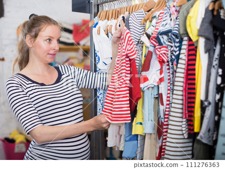 Pregnant woman in striped tunic chooses shirt in clothing shop Pregnant woman in striped tunic chooses shirt in clothing shop 111276363