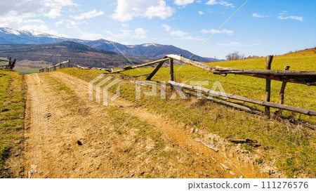 country road through the hill with grassy meadow behind the wooden fence. carpathian mountain ridge with snow capped tops in the distance. beauty of ukrainian countryside in spring 111276576