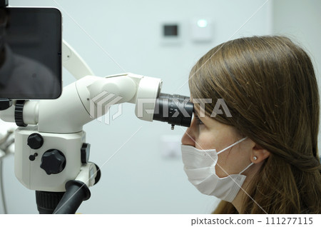 Brunette woman dentist in a white coat and a medical protective mask works with a modern microscope in the office. Operations in the dental clinic. Brunette woman dentist in a white coat and a medical protective mask works with a modern microscope in the office. Operations in the dental clinic. 111277115