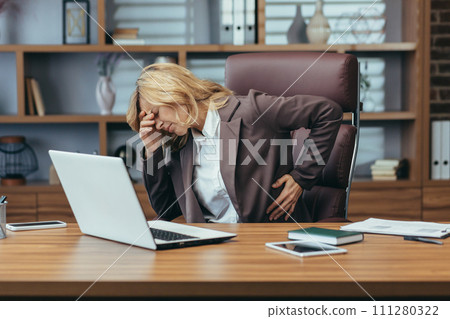 Senior business woman in a suit sitting at a desk in the office holding her hand on her back and head and suffering from severe pain. Needs rest and medical assistance. 111280322
