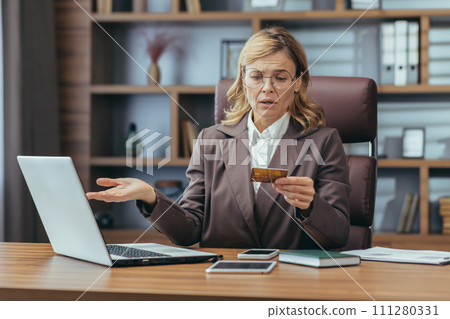 Shocked and worried senior business woman sitting at the desk in the office and looking at the credit card she is holding with her arms wide open. Shocked and worried senior business woman sitting at the desk in the office and looking at the credit card she is holding with her arms wide open. 111280331