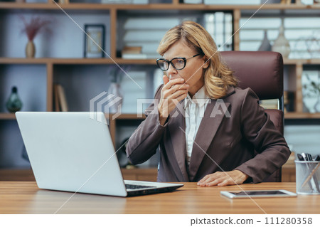 Shocked senior business woman sitting in the office at the desk and looking worriedly at the laptop screen. Problems with accounts, received bad news in the mail, reads the message. Shocked senior business woman sitting in the office at the desk and looking worriedly at the laptop screen. Problems with accounts, received bad news in the mail, reads the message. 111280358