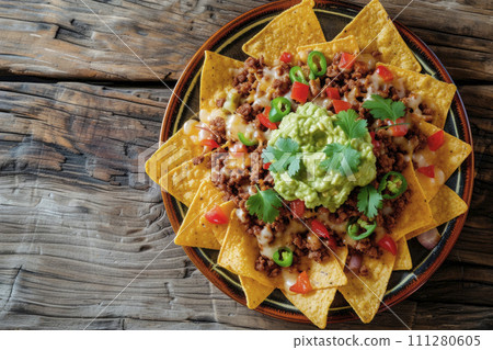 Mexican nachos with beef, guacamole, cheese sauce, peppers, tomato and onion in plate on wooden table. Top view. Copy space Mexican nachos with beef, guacamole, cheese sauce, peppers, tomato and onion in plate on wooden table. Top view. Copy space 111280605