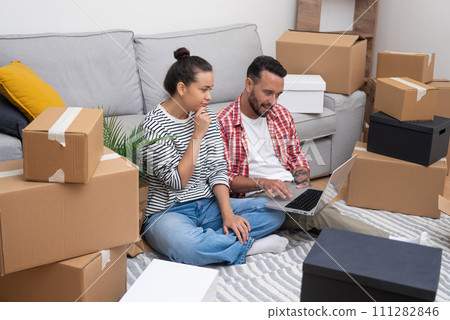 Young spouse sits on floor among newly unpacked boxes of things thoughtfully selecting from web site comfortable things for living room and kitchen of new home 111282846