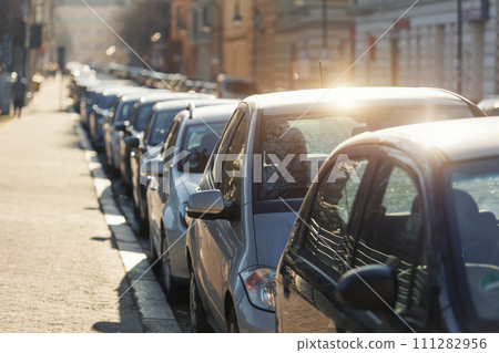 Scenic sunlit view row parked cars on busy city street in european city. Sidewalk parallel side parking full of vehicles traffic jam in downtown on warm sunset evening or sunrise morning 111282956