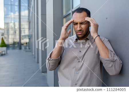 Sick diverse male suffering from severe headache and rubbing forehead while standing by wall outdoors. Tired exhausted man feeling unexpected dizziness and struggling with high blood pressure. Sick diverse male suffering from severe headache and rubbing forehead while standing by wall outdoors. Tired exhausted man feeling unexpected dizziness and struggling with high blood pressure. 111284005