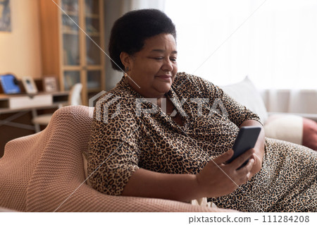 Side view portrait of smiling Black senior woman using smartphone relaxing on couch in cozy home copy space 111284208