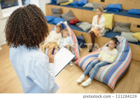 High angle view at Black young woman holding clipboard and taking notes while working with group of children as teacher or therapistwith copy space High angle view at Black young woman holding clipboard and taking notes while working with group of children as teacher or therapistwith copy space 111284229