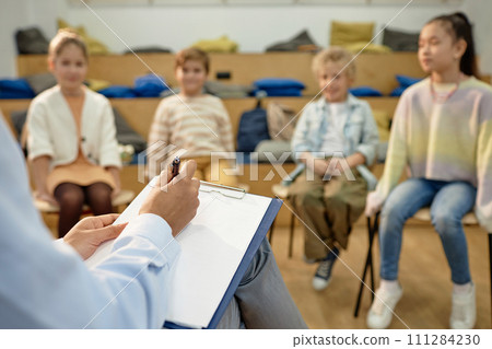 Close up of teacher or therapist holding clipboard and taking notes while working with group of childrenwith copy space Close up of teacher or therapist holding clipboard and taking notes while working with group of childrenwith copy space 111284230
