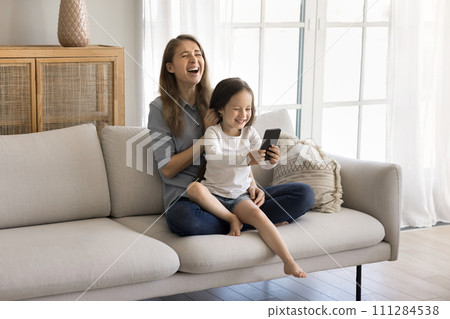 Kid and mom laugh sit on sofa with smartphone 111284538