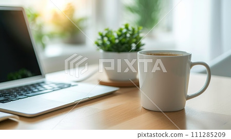 A white mug next to a laptop on a wooden table, illuminated by warm sunlight 111285209