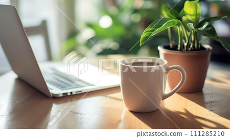 A white mug next to a laptop on a wooden table, illuminated by warm sunlight 111285210
