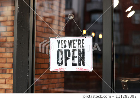 Inscription Yes we're open metal plate with black and white sign on glass door store, cafe, beautystore, barbershop after coronavirus lockdown quarantine. business reopen again 111285392