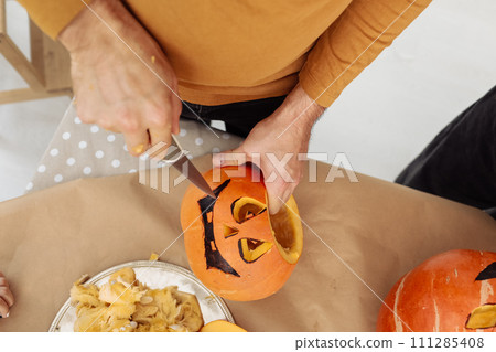 Close up of man hands carving Jack O Lattern from ripe orange pumpkin with knife on her wooden kitchen table. male preparing all hallows eve Halloween party decorations. Background, copy space. 111285408