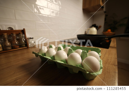 White chicken eggs in green cardboard egg tray on tabletop at modern stylish kitchen. Black frying wok pan on induction stove. Delicious healthy simple breakfast meal. Natural egg protein to the diet 111285455