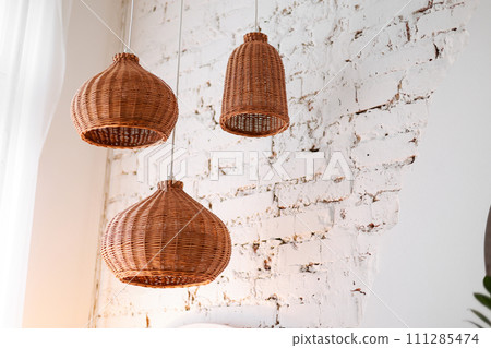three rattan chandeliers different forms on white brick wall background. Straw lampshade in cozy living room. Eco-friendly interior design using natural materials. Scandinavian interior. copy space three rattan chandeliers different forms on white brick wall background. Straw lampshade in cozy living room. Eco-friendly interior design using natural materials. Scandinavian interior. copy space 111285474