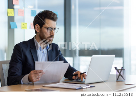 Serious and focused financier accountant on paper work inside office, mature man using calculator and laptop for calculating reports and summarizing accounts, businessman at work in suit. 111285518