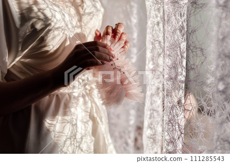 Bride morning preparation. Beautiful bride in white wedding negligee on the background of large bedroom window. High quality photo Bride morning preparation. Beautiful bride in white wedding negligee on the background of large bedroom window. High quality photo 111285543
