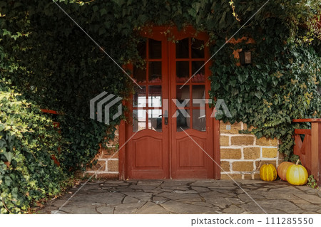 Exterior of building facade with retro wooden brown entrance door with glass and overgrown green ivy on bricks wall, stone path. Autumn season with pumpkins. Thanksgiving holiday. Halloween Exterior of building facade with retro wooden brown entrance door with glass and overgrown green ivy on bricks wall, stone path. Autumn season with pumpkins. Thanksgiving holiday. Halloween 111285550