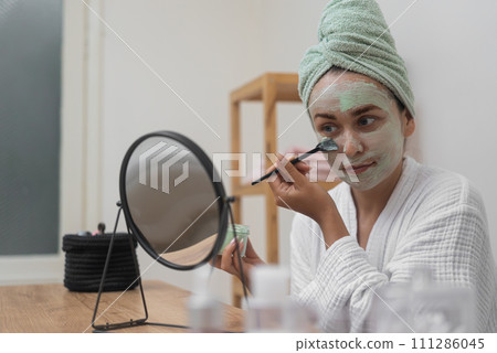 Skincare Retreat: A bathrobe-clad beauty uses a brush to apply a clay mask, admiring her reflection during her organic facial treatment. Skincare Retreat: A bathrobe-clad beauty uses a brush to apply a clay mask, admiring her reflection during her organic facial treatment. 111286045
