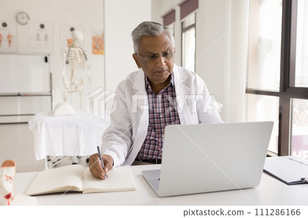 Mature Indian doctor man in uniform coat working at laptop Mature Indian doctor man in uniform coat working at laptop 111286166