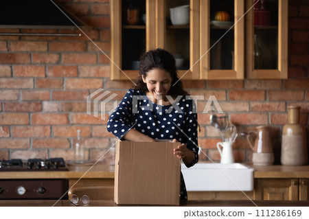 Excited woman unpacking carton box in kitchen, looking inside 111286169