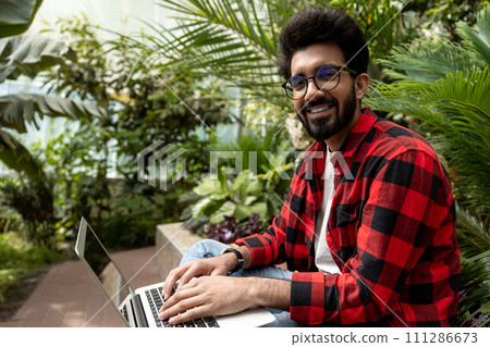 Hindu man in checkered shirt working on laptop in the garden Hindu man in checkered shirt working on laptop in the garden 111286673