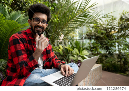 Hindu man in checkered shirt working on laptop in the garden Hindu man in checkered shirt working on laptop in the garden 111286674