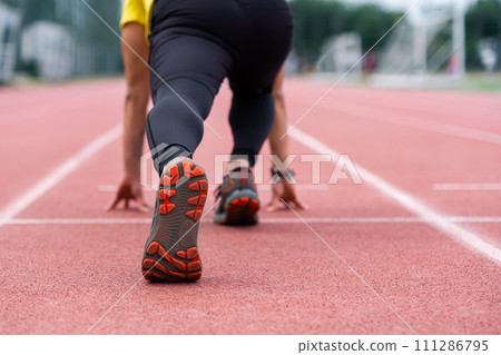Runner standing in low start position ready to run along red rubberized track on urban ground professional sportsman exercising at outdoor stadium closeup Runner standing in low start position ready to run along red rubberized track on urban ground professional sportsman exercising at outdoor stadium closeup 111286795