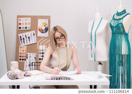 Thoughtful woman with glasses studies clothes sketch at desk in fashion atelier green macrame dress on mannequin near designer workplace in sewing shop Thoughtful woman with glasses studies clothes sketch at desk in fashion atelier green macrame dress on mannequin near designer workplace in sewing shop 111286814