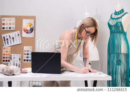 Woman designer marks notes at desk with laptop in dressmaking workshop tailor creates women clothes by mannequin with macrame dress in fashion atelier Woman designer marks notes at desk with laptop in dressmaking workshop tailor creates women clothes by mannequin with macrame dress in fashion atelier 111286815