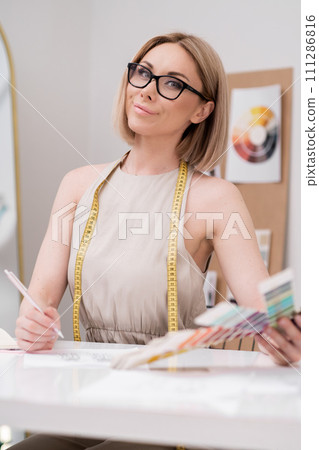 Smiling lady designer with glasses and color swatch draws at workplace in dressmaking atelier professional tailor creates apparel sketches in sewing studio dressmaker job Smiling lady designer with glasses and color swatch draws at workplace in dressmaking atelier professional tailor creates apparel sketches in sewing studio dressmaker job 111286816
