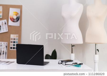 Clothes designer workplace with laptop in dressmaking shop notebook computer on desk in fashion atelier manager table and mannequins in empty sewing studio Clothes designer workplace with laptop in dressmaking shop notebook computer on desk in fashion atelier manager table and mannequins in empty sewing studio 111286820