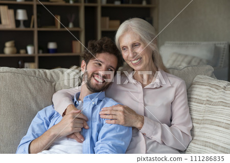 Mature woman hugging adult son resting on sofa at home Mature woman hugging adult son resting on sofa at home 111286835