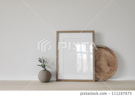 Blank vertical picture frame mockup, poster display. Ball vase with olive tree branch. Round wooden tray on table, desk. Minimal rustic home. Scandinavian interior. White wall background. 111287875