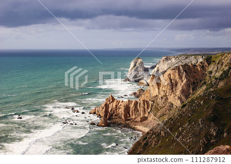 Beautiful steep coast, rocks at Praia da Ursa beach, Portugal. Dramatical sunset with stormy clouds sky at Cape Roca. Turqoise waters, waves of Atlantic ocean. Travel Europe. Cabo da Roca. Sintra 111287912