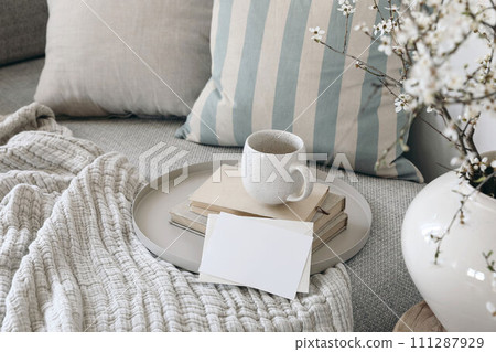 Spring breakfast scene. Blank greeting card mockup. Cup of coffee, tea on books. Round beige tray. Blossoming cherry plum tree branches in ceramic vase. Cozy linen sofa, cushions. Easter still life. 111287929