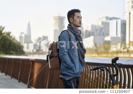 young asian adult man with backpack standing by his bike looking at city skyline in riverside park young asian adult man with backpack standing by his bike looking at city skyline in riverside park 111288907