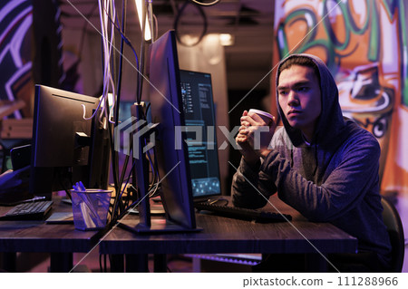 Hacker drinking coffee to go while doing illegal criminal activity on computer. Young man in hood holding beverage mug and doing ddos attack on website in abandoned warehouse 111288966