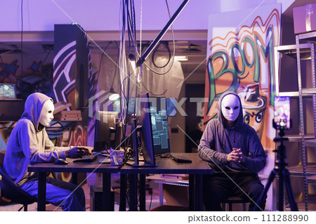 Internet scammers in anonymous masks threatening victim online on smartphone. Hackers recording ransom message video on mobile phone while working together in abandoned warehouse 111288990