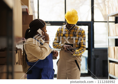Warehouse worker checking parcel in tablet and manager answering supervisor call. African american woman talking on landline phone and assistant using inventory software in storehouse 111288992
